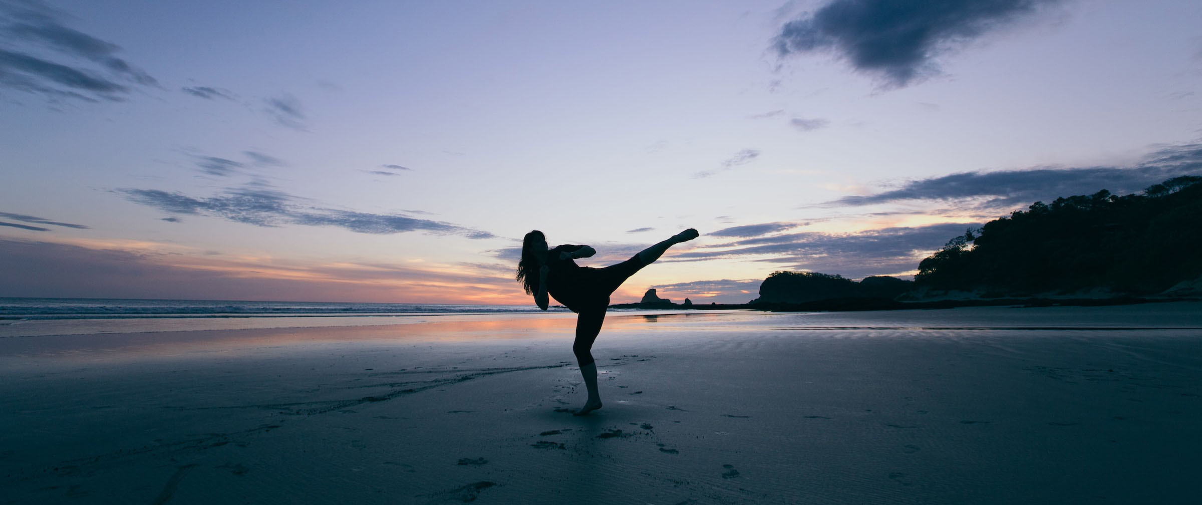 Eine sportliche Frau führt im Sonnenuntergang am Strand einen Karate-Kick aus. 